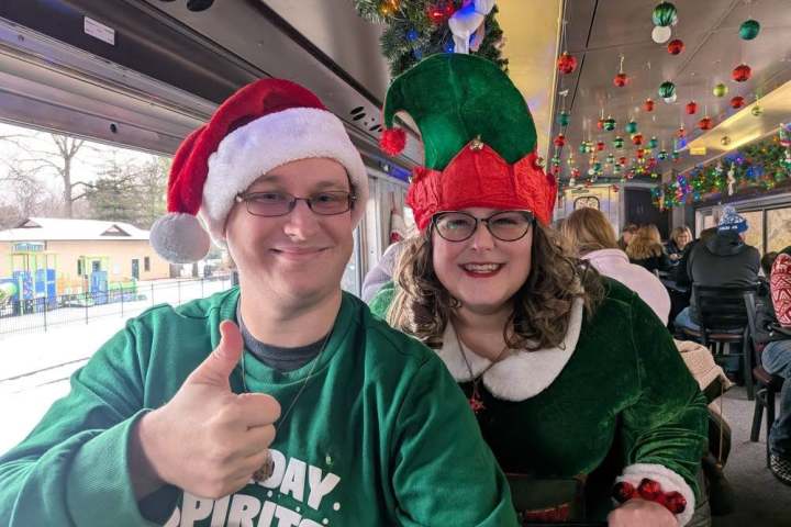 Two people in festive attire, one in Santa hat, the other in elf costume, smiling on a decorated train.