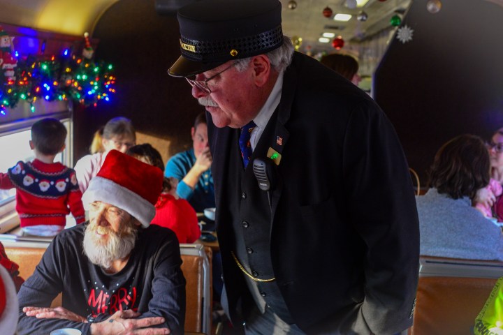 Train conductor talks to elderly man with Santa hat on holiday-decorated train.