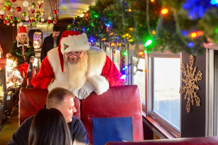 Santa leans over train seat, talking with passengers on a decorated holiday train.