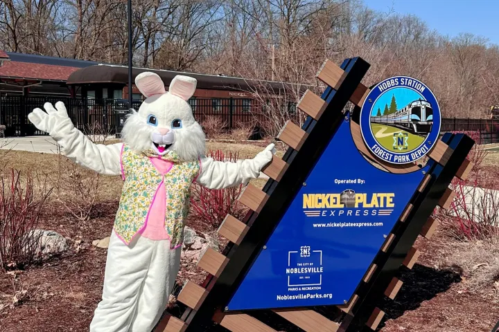 Person in Easter bunny costume stands beside a Hobbs Station sign on a sunny day.