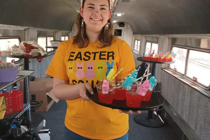 Person in bunny ears and Easter shirt holds tray with colorful drinks inside a train car.