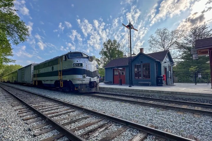 Vintage train at a station with a small building, surrounded by trees and a cloudy sky.