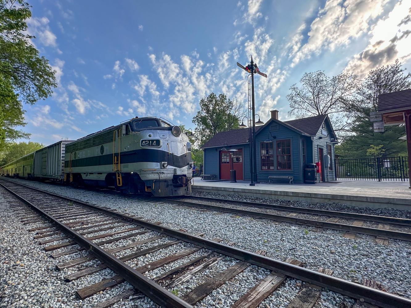Vintage train at a small station with a signal pole and cloudy sky.