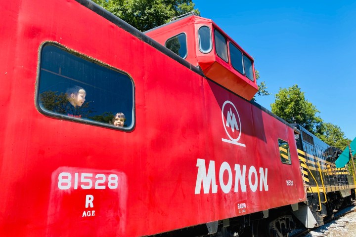 Red Monon train car with two children looking out the window, green trees and blue sky in the background.