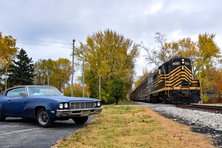 Vintage blue car parked near train tracks with train passing by on an overcast day.