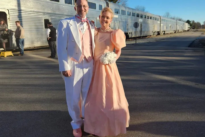 Couple in formal attire posing by a train under a clear blue sky.
