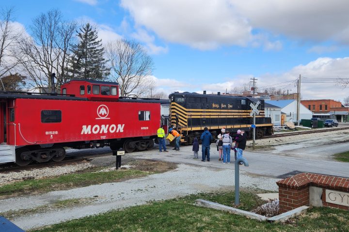 Red caboose and black train on tracks with people nearby under a blue sky.