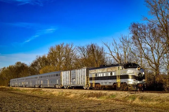 Vintage train traveling on tracks through a rural landscape with trees and clear blue sky.