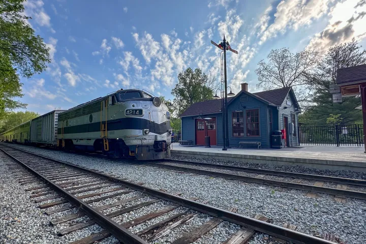Train at small station with vintage signal, trees and cloudy sky in background.