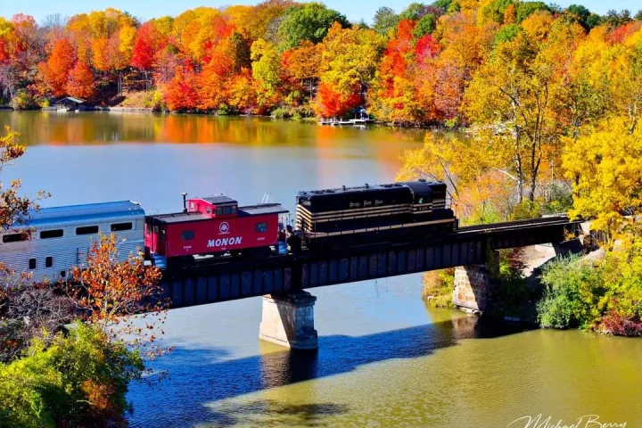 Train crosses bridge over river with colorful autumn trees in the background.