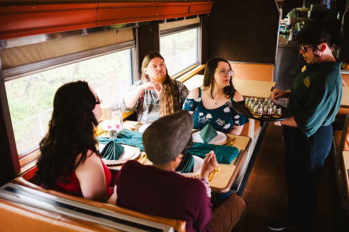 Four people seated in a train dining car being served drinks by a waiter.