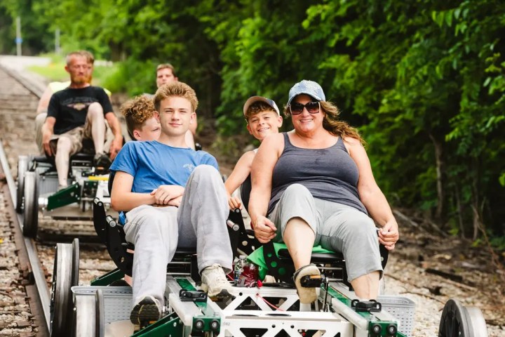 Group of people riding pedal-powered rail carts on tracks surrounded by trees.