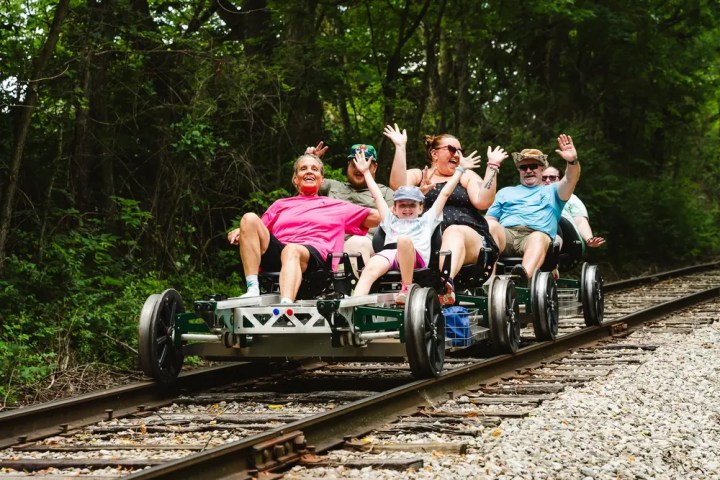 Group of people riding a railbike on train tracks in the woods, smiling and raising their hands.