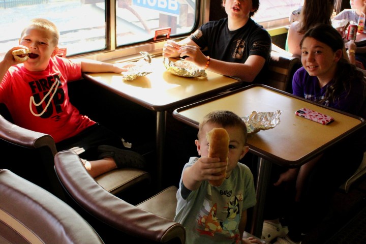 Children sitting at a table on a train, one holding a hot dog.