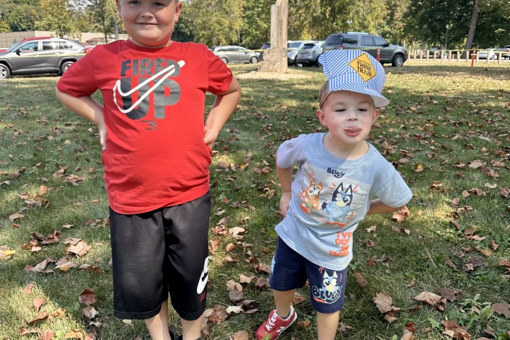 Two children wearing striped train hats standing outdoors on grass.