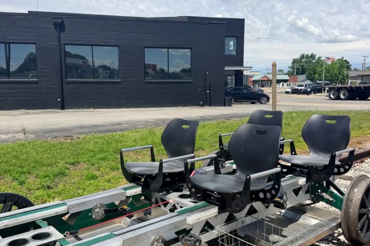 Rail cart on tracks with four black seats, building in background.