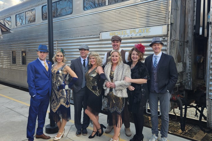 Group in vintage attire poses by a train on a sunny day.
