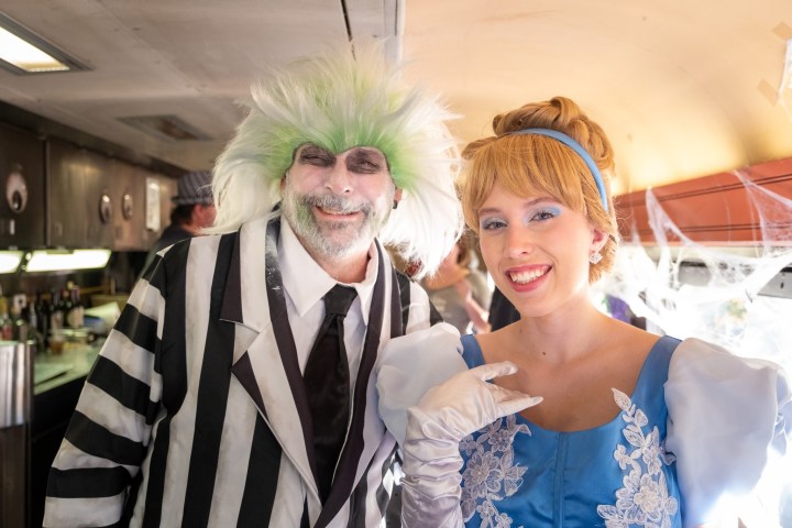 Man in black-and-white costume with green hair and woman in blue dress smiling in a decorated setting.