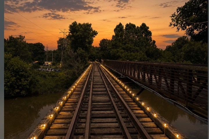 Railroad track over water at sunset, lit by lights, with trees and bridge in background.